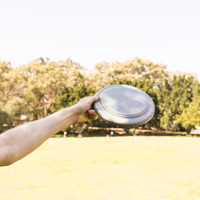 A hand grasps a grey frisbee against a blurred green park background, featuring trees and grass.