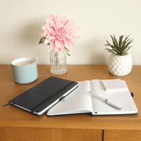 A Pineapple Leather Bound JournalBook in black, opened with a pen, alongside a flower vase and potted plant on a wooden surface.