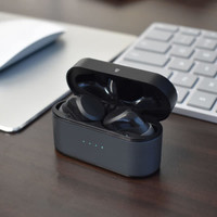 A black charging case with wireless earbuds inside, placed on a wooden surface beside a keyboard and a laptop.