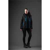 A woman in a black jacket with blue accents stands against a dark background, wearing black leggings and boots.