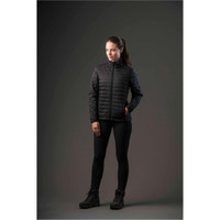 A woman models a black puffy jacket, paired with black leggings and boots, set against a dark background.