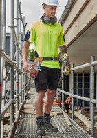 A man wearing a high-visibility yellow polo shirt and black cargo shorts, carrying tools on a construction site.