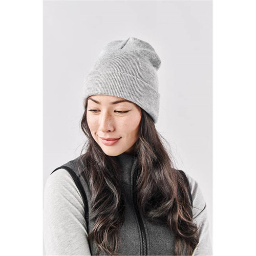 A grey knit beanie worn by a woman with long, wavy hair, paired with a dark vest. It features a logo. A grey knit beanie worn by a woman with long, wavy hair, paired with a dark vest. It features a logo.
