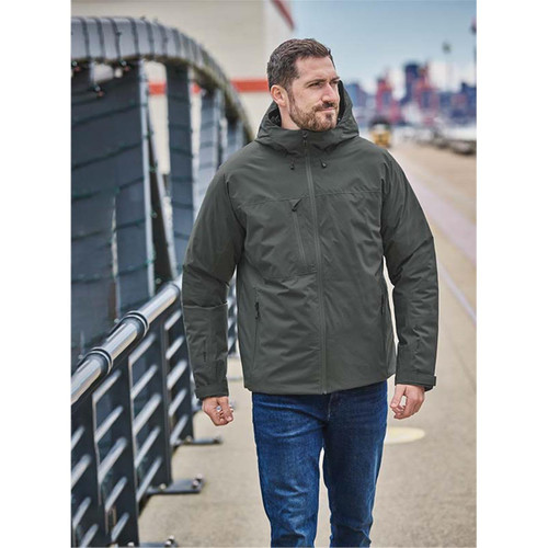 A man wearing a dark green thermal shell jacket, walking outdoors along a paved path. The jacket features a hood. A man wearing a dark green thermal shell jacket, walking outdoors along a paved path. The jacket features a hood.