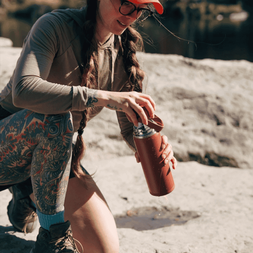 A woman with tattoos and a red hat holding a red water bottle kneeling on a light grey rock