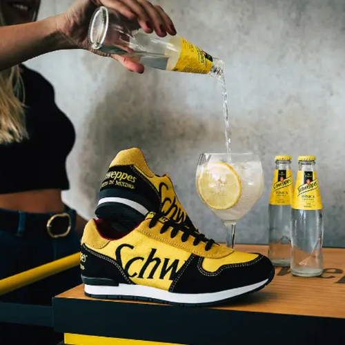 A hand pours Schweppes tonic water into a glass with a lemon slice, beside custom yellow and black sneakers featuring the brand's logo. A hand pours Schweppes tonic water into a glass with a lemon slice, beside custom yellow and black sneakers featuring the brand's logo.