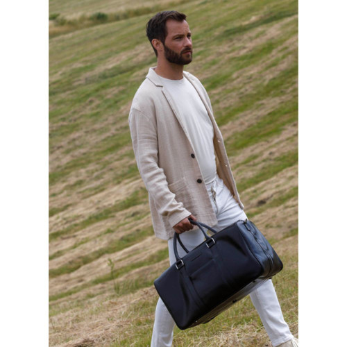 Man in a beige blazer holding a black leather duffel bag outdoors.
