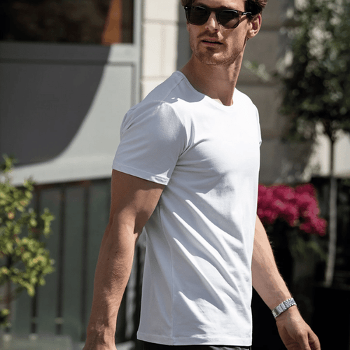 A man wearing a white T-shirt and sunglasses, walking outdoors with a bag in hand.