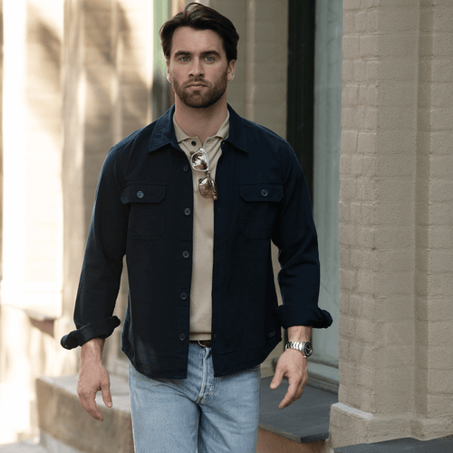 A unisex shirt in navy with buttons, worn over a beige top, paired with light jeans. The model is walking outdoors.