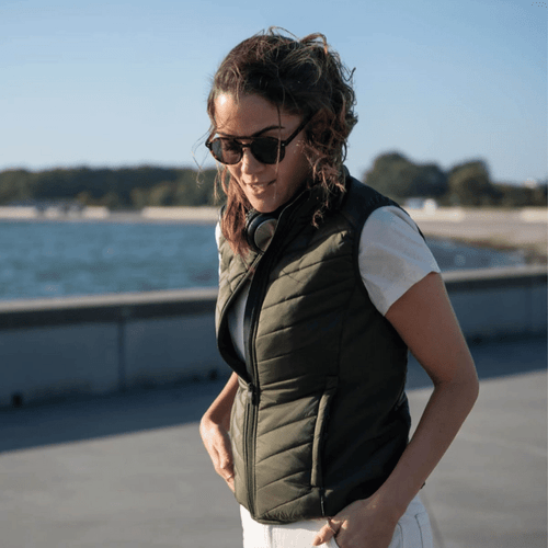 A woman wearing an olive green vest over a white shirt, standing by a waterfront, with sunglasses on.