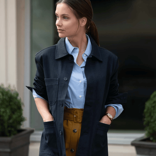 A woman wearing a dark navy over shirt with a light blue top and light brown pants, both hands in the side pockets of the shirt.