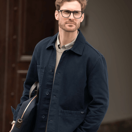 A man wearing a navy unisex shirt, paired with a light grey sweater and green trousers, holding a black bag.