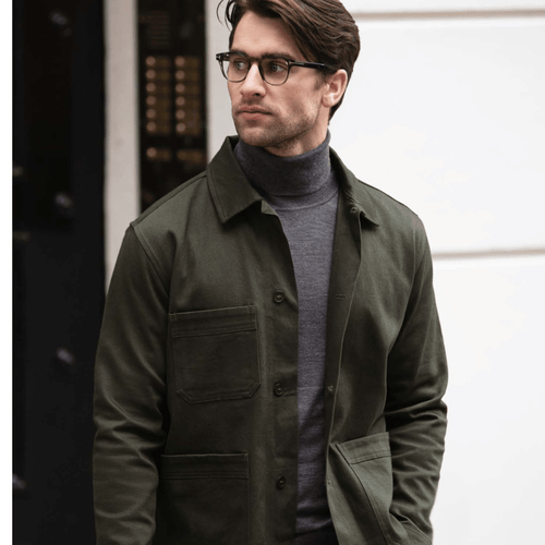 A man wearing a dark green unsiex shirt, paired with a grey turtleneck jumper standing outside in the sun in front of a light coloured house wall.