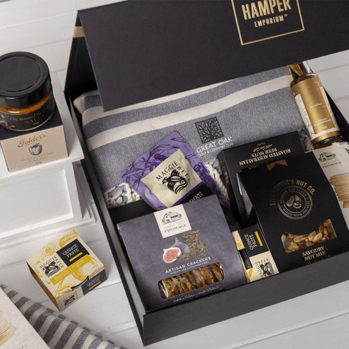 A gift pack featuring a grey blanket, gourmet snacks, nut mix, crackers, and a jar of preserve, all in a black box.
