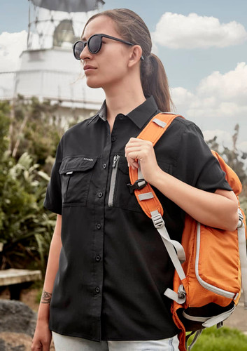 A woman in a black short sleeve outdoor shirt carries an orange backpack, standing near a lighthouse.