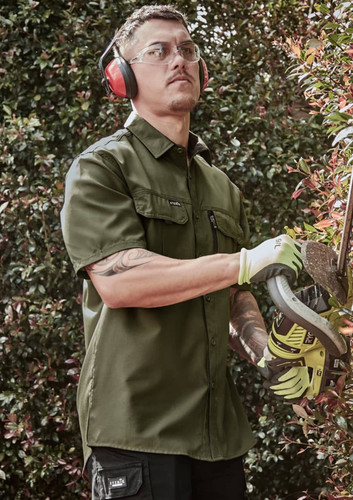 A man wearing an olive green short sleeve button-up shirt, safety glasses, and ear protection, using a power tool.