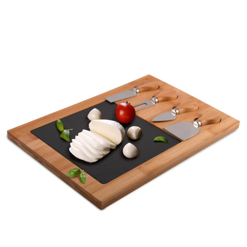 A bamboo cheese board with a black slate surface, featuring cheese, a tomato, and three stainless steel cheese knives.