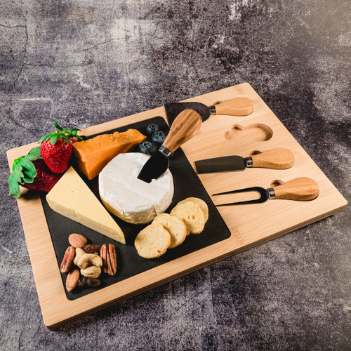 A bamboo cheese board featuring a slate surface, assorted cheeses, fruits, nuts, and cheese knives.