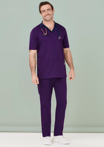 A man wears purple scrub pants with a matching scrub top and stethoscope, set against a pale background.