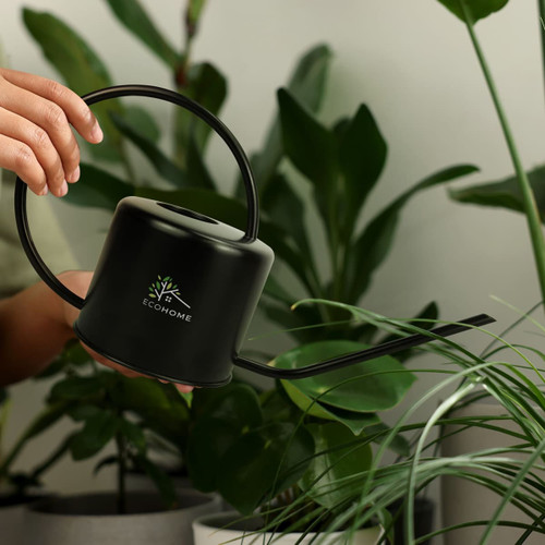 A black watering can with a curved handle, set against lush green plants. It features a logo.