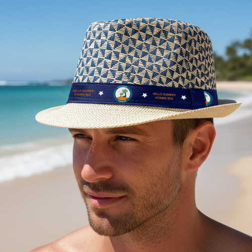 A straw woven top hat with a patterned design and a blue band featuring a logo, set against a beach background.