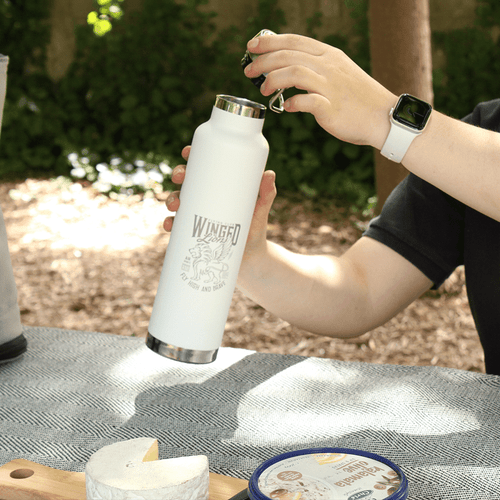 A drink bottle in white with a silver lid, featuring a logo, held by a person outdoors. A drink bottle in white with a silver lid, featuring a logo, held by a person outdoors.