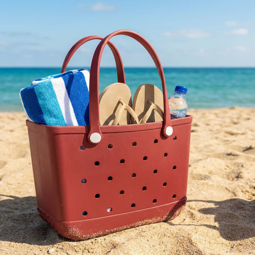 A maroon beach bag with white handles, containing a blue and white towel, flip-flops, and a drink bottle on the sand by the sea.