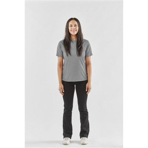 A woman stands smiling, wearing a grey women's t-shirt paired with black pants and white shoes. The shirt has a logo.