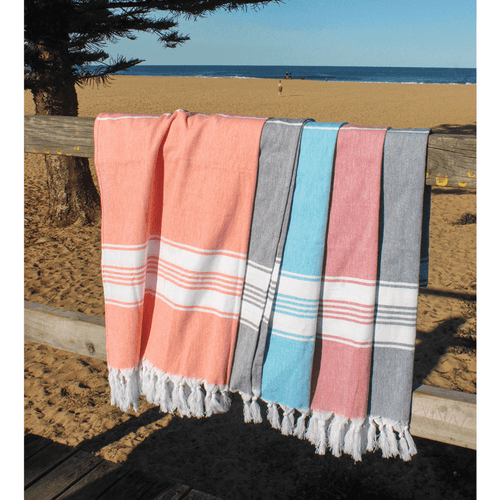 A collection of four Turkish towels in coral, blue, grey, and beige draped over a wooden railing against a beach backdrop.
