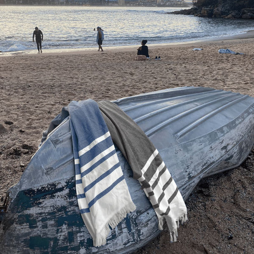 A grey and blue striped towel draped over a weathered boat on a sandy beach, with people in the background. A grey and blue striped towel draped over a weathered boat on a sandy beach, with people in the background.