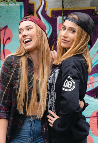 Two women wearing organic brushed heavy cotton/mesh back caps, one in maroon and the other in black, smiling against a graffiti background.