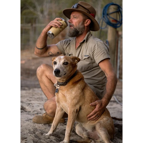 Lifestyle image of a person outdoors holding a beige insulated fridgy® tumbler with a clear lid, accompanied by a dog in a rugged natural setting.