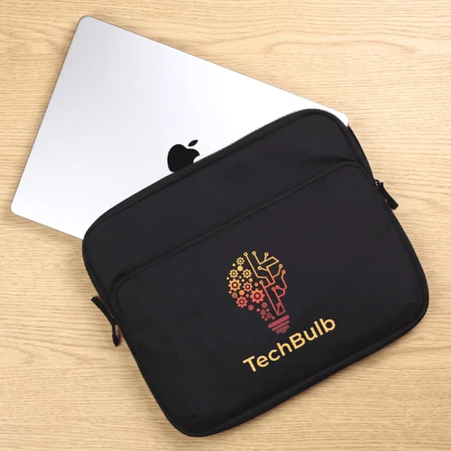 A black laptop sleeve featuring a logo design, with a silver laptop partially visible behind it on a wooden surface.