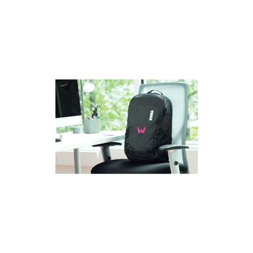 A black laptop backpack with a logo, positioned on a chair beside a desk with a computer and stationary.