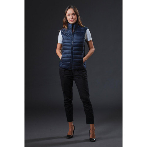 A navy women's puffer vest is displayed on a model, featuring a sleek design and a logo. The model wears black pants and heels. A navy women's puffer vest is displayed on a model, featuring a sleek design and a logo. The model wears black pants and heels.