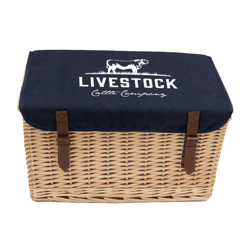 A wicker picnic basket with a navy blue canvas lid, featuring branding and leather straps for closure.
