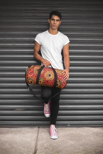 A duffle bag in red and black with a floral pattern is held by a person in a fitted white t-shirt and pink sneakers.