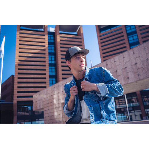 A young man wearing a grey and black trucker cap, styled with a denim jacket, standing in an urban environment. A young man wearing a grey and black trucker cap, styled with a denim jacket, standing in an urban environment.