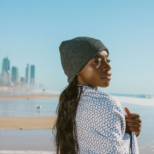 Person wearing a beanie and patterned jacket on a sunny beach with city skyline in background.