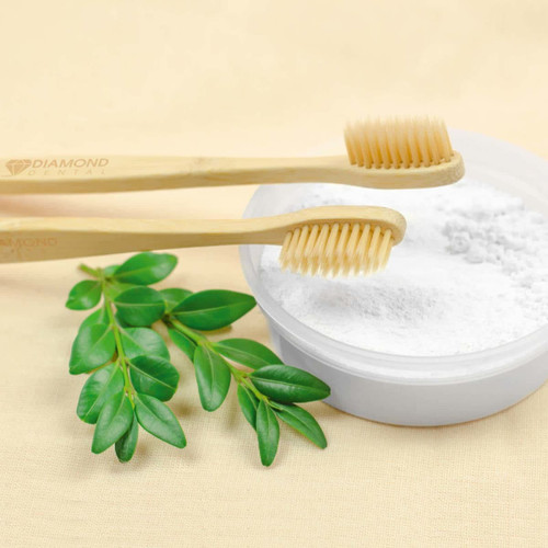A bamboo toothbrush with soft bristles is placed next to a bowl of white powder, accompanied by green leaves. A bamboo toothbrush with soft bristles is placed next to a bowl of white powder, accompanied by green leaves.