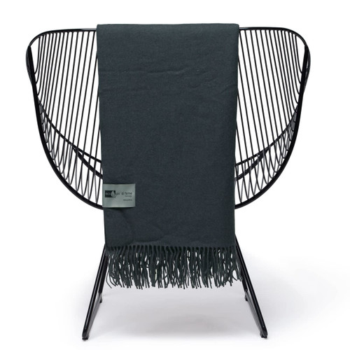 A dark green Merino wool blanket with fringed edges, displayed on a modern wire chair, featuring a branding label. A dark green Merino wool blanket with fringed edges, displayed on a modern wire chair, featuring a branding label.