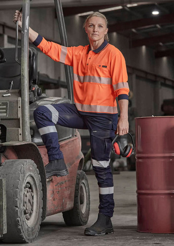 A woman in an orange high-visibility shirt and dark cargo pants leans against a forklift, wearing safety gear.