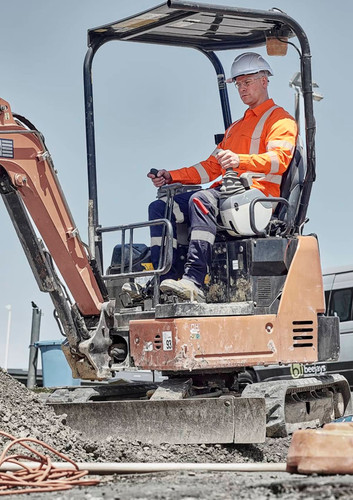 A worker in a high-visibility orange shirt and white hard hat operates a small excavator on a construction site. A worker in a high-visibility orange shirt and white hard hat operates a small excavator on a construction site.