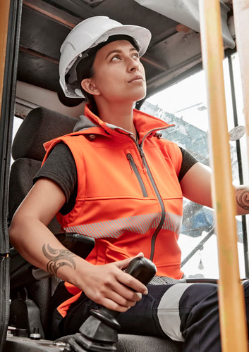 A woman wearing an orange softshell jacket with reflective strips, seated in a construction vehicle, wearing a hard hat. A woman wearing an orange softshell jacket with reflective strips, seated in a construction vehicle, wearing a hard hat.