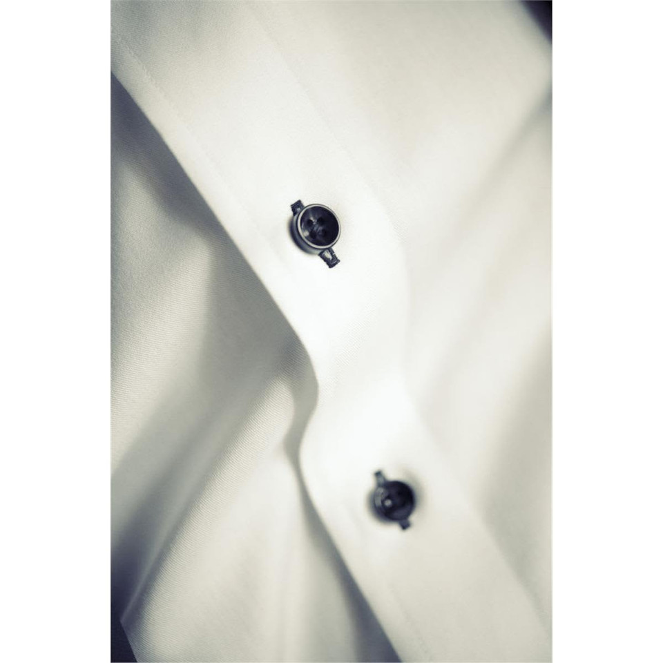 A close-up of a white button-up shirt featuring black buttons and a subtle texture, with a branding logo visible.