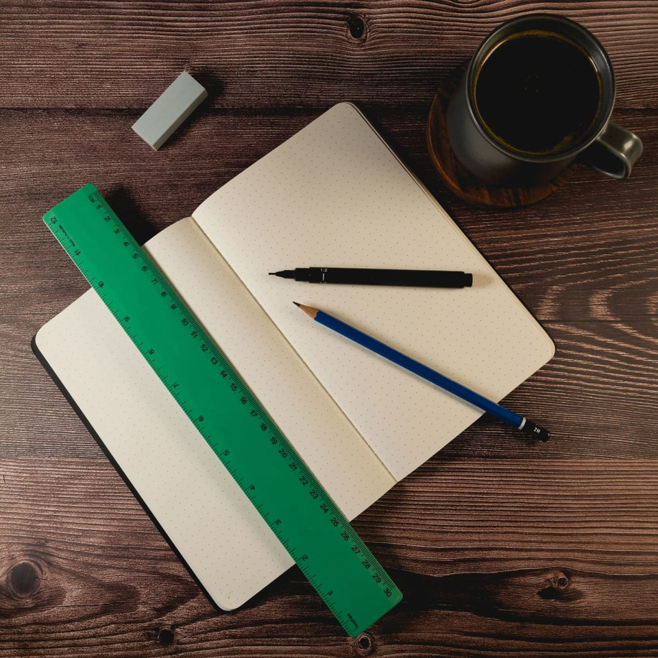 A green recycled plastic ruler lies on an open notebook, accompanied by a black pen, a blue pencil, and a coffee mug.