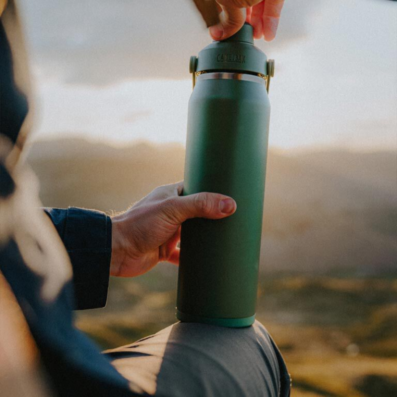 Person holding a green Camelbak stainless steel water bottle against a sunset backdrop, symbolizing hydration and outdoor adventures.