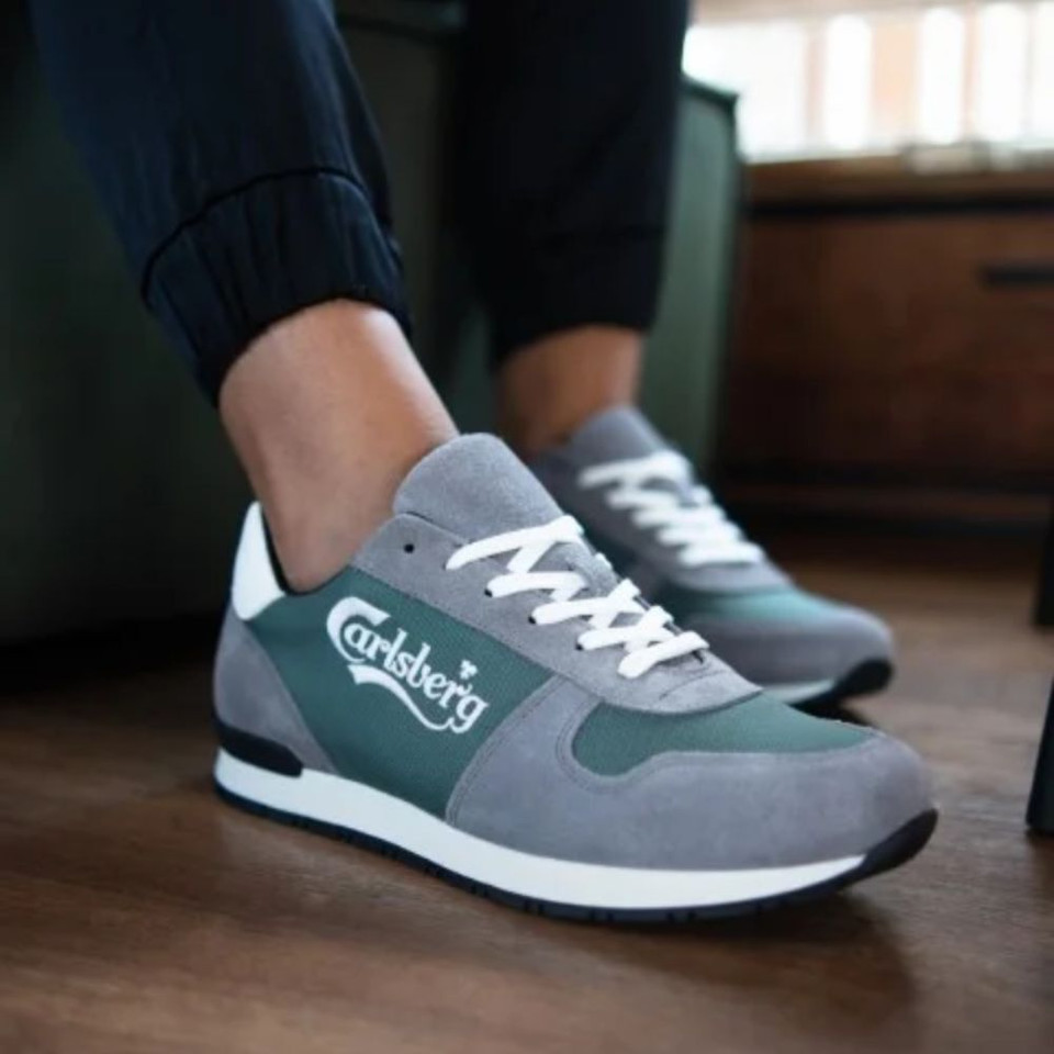 Custom carlsberg sneakers in grey and green with white laces worn by feet. The legs are stuck in black pants