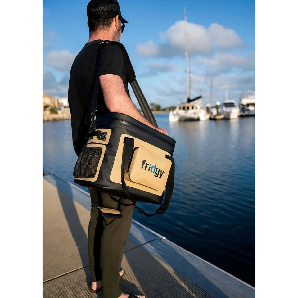 Lifestyle image of a black and tan Fridgy insulated cooler bag with shoulder strap and external storage pockets, carried beside a marina waterfront.