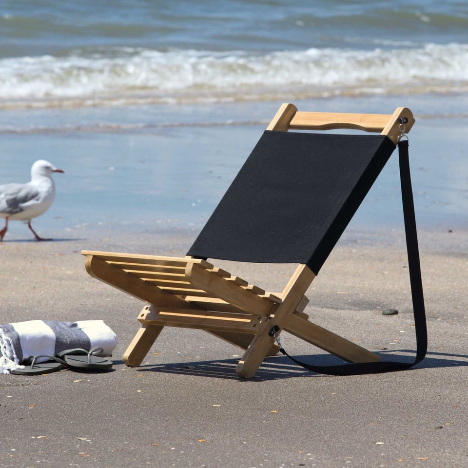A wooden beach chair with a black fabric seat sits on the sand near the water. Two rolled towels are beside it.